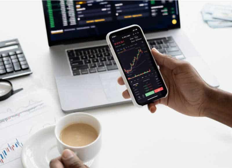 Hand held smartphone and laptop on desk showing stock charts